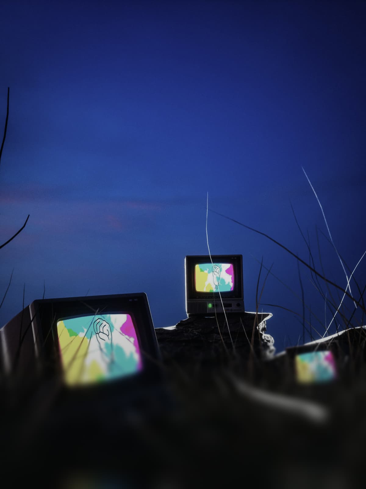 Mehrere alte Röhrenfernseher stehen nachts zwischen hohem Gras und zeigen farbenfrohe Bilder.