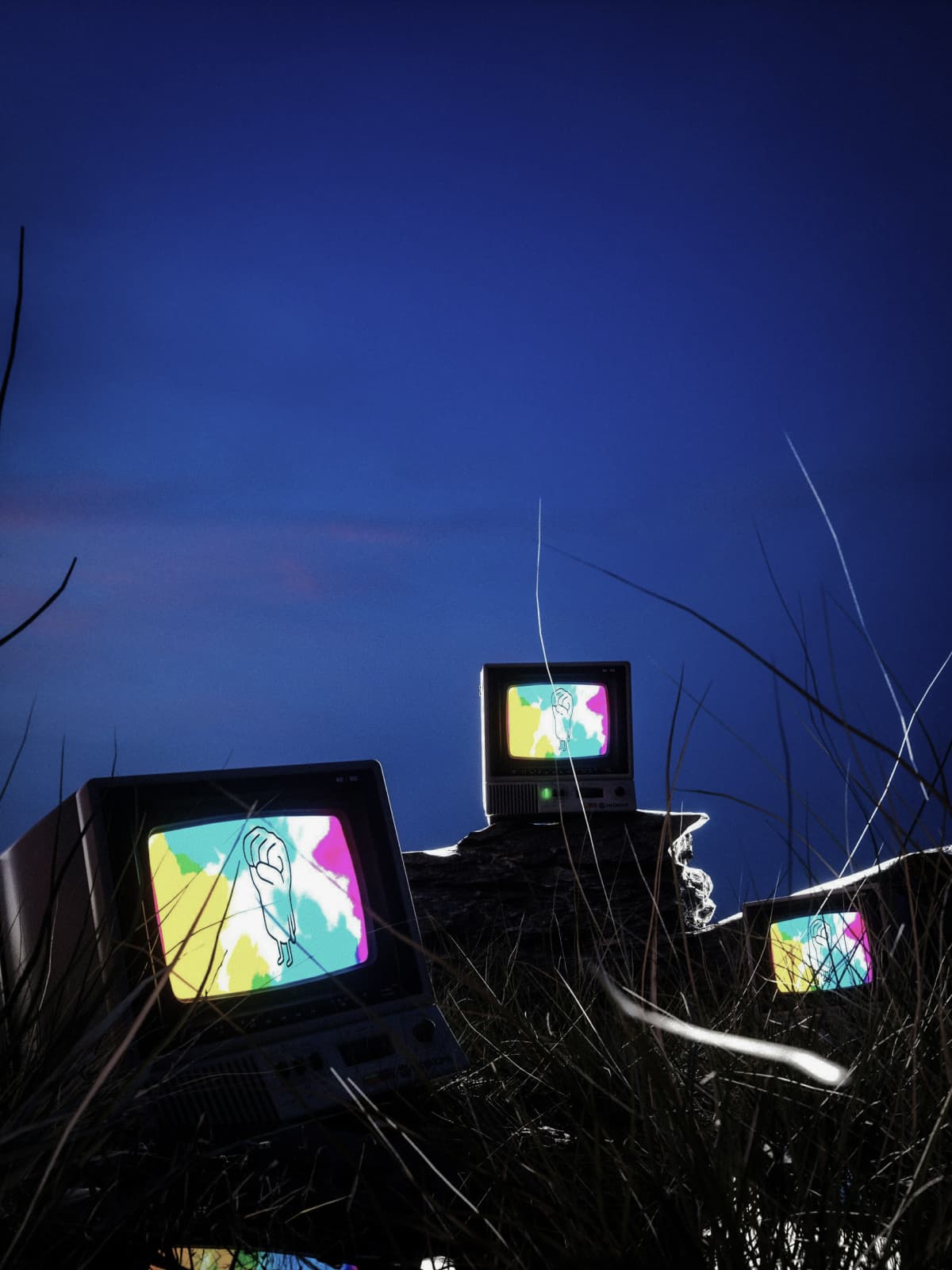 Mehrere alte Röhrenfernseher stehen nachts zwischen hohem Gras und zeigen farbenfrohe Bilder.